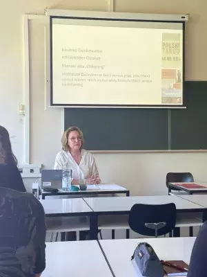 prof. Szymańska podczas wykładu, siedzi przy biurku, za nią wyświetlana jest prezentacja